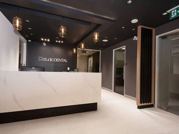 A modern dental office reception area featuring a sleek white desk, dark walls, and contemporary lighting fixtures. The space is well-lit, inviting, and designed for patient comfort. The 123Dentist Smiley adds a friendly touch to the welcoming atmosphere.