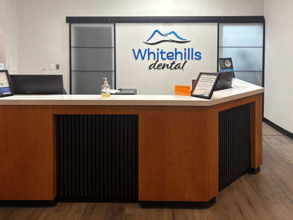 Une zone de réception de cabinet dentaire avec un bureau d'accueil moderne en bois, un logo « Whitehills Dental » au mur et des présentoirs d'information. L'espace est bien éclairé avec une esthétique propre et accueillante.