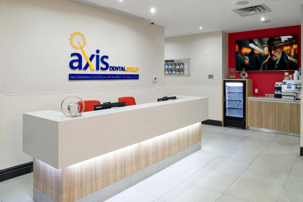 A modern dental office reception area featuring a sleek white and wood front desk with red chairs. The wall displays the "axis dental" logo, and a television screens a dark image. A beverage fridge is visible to the right.