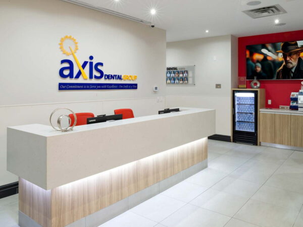 A modern dental office reception area featuring a sleek white and wood front desk with red chairs. The wall displays the "axis dental" logo, and a television screens a dark image. A beverage fridge is visible to the right.