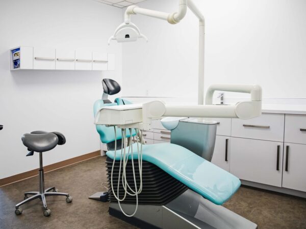 A modern dental clinic featuring a teal dental chair, overhead light, and a dental assistant's stool. Clean white cabinetry is visible in the background, emphasizing a professional and inviting environment.
