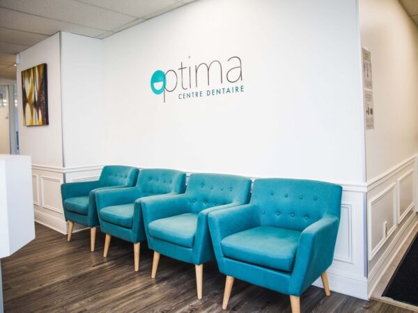A modern dental clinic reception area featuring a branded wall sign for "Optima Centre Dentaire." The space includes several teal chairs arranged against a white wall, creating a welcoming atmosphere.