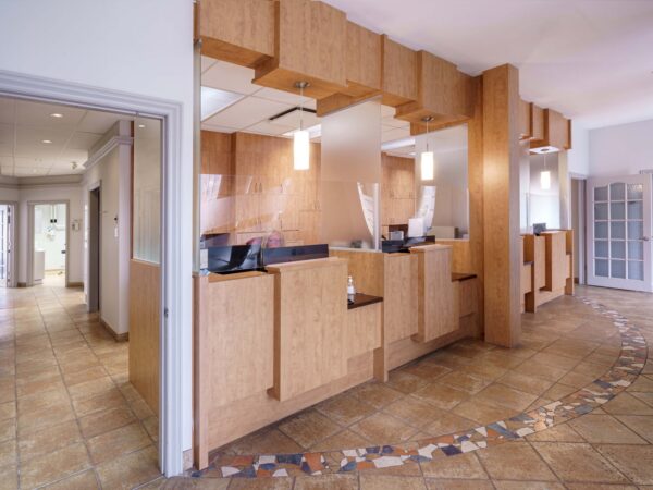 A modern, well-lit reception area featuring a wooden front desk and tiled flooring. The space is inviting with natural light coming through windows, leading to a hallway in the background.