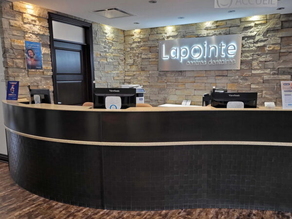 A modern reception area with a curved black counter, stone walls, and the name "Lapointe" illuminated in light. The space is clean and inviting, featuring check-in stations and informational displays.