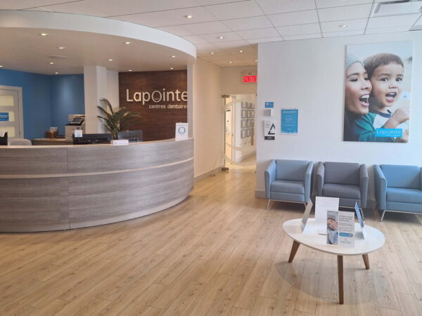 A modern reception area featuring a curved wooden desk with a plant. Light blue walls complement gray seating and a coffee table. A large poster of smiling people is displayed on the wall, creating an inviting atmosphere.