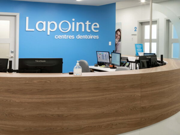 A modern dental office reception area featuring a curved wooden desk, multiple computer stations, and a blue wall with the sign "Lapointe centres dentaires." The space is bright and welcoming, designed for patient comfort and efficient service.