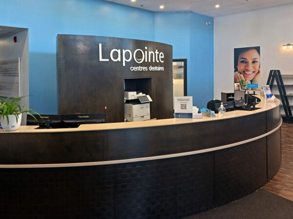 A friendly dental office reception area featuring a curved front desk with the name "Lapointe" prominently displayed. The walls are painted light blue, and the space includes promotional materials and a smiling image of a woman, creating a welcoming atmosphere for patients. The 123Dentist Smiley adds a cheerful touch.