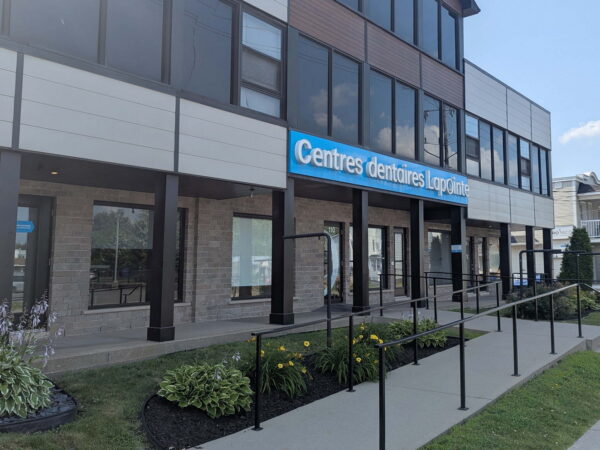 A modern dental clinic building with large windows and a blue sign that reads "Centres dentaires Longueuil." The entrance features a ramp, bordered by green landscaping, in a well-lit, urban setting.