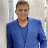 A smiling individual stands confidently in a bright blue suit against a softly colored background, exuding warmth and professionalism.