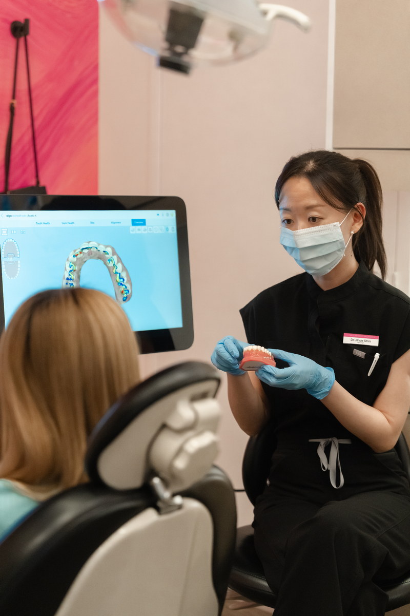 A dental professional wearing a mask and gloves explains a treatment to a patient seated in a dental chair, with a digital monitor displaying an orthodontic model in the background. The scene conveys a friendly, informative atmosphere in a dental office.
