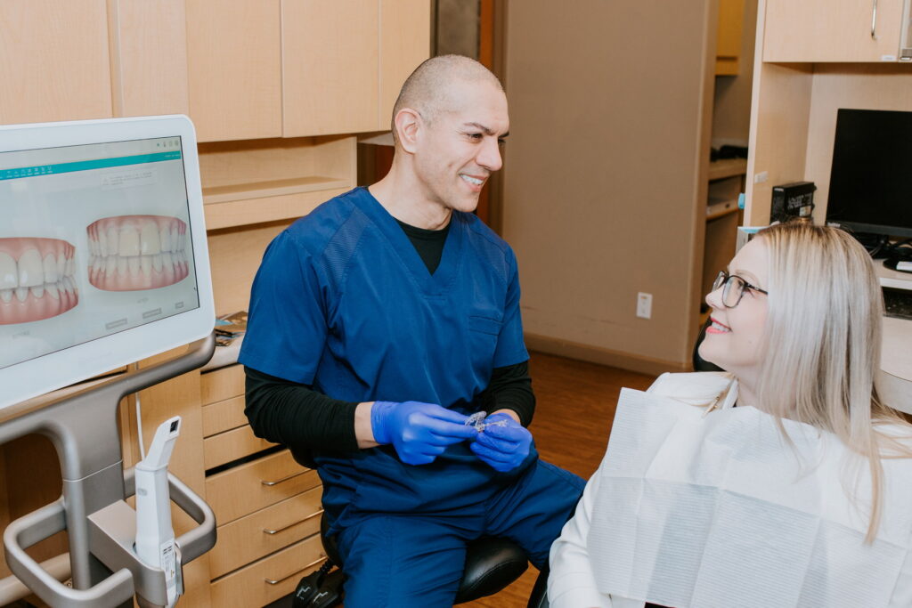 A dentist in blue scrubs smiles while interacting with a patient who is seated in a dental chair. The patient, wearing a bib, appears relaxed as they engage in conversation, with dental preparation tools and a computer screen displaying dental images in the background.