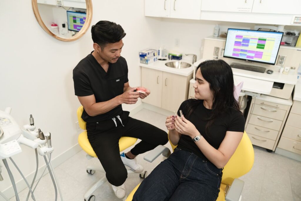 A dental professional and a patient engage in conversation inside a modern dental office. The professional is seated on a yellow chair, demonstrating a dental tool to the patient, who appears engaged and attentive. Background features include a computer and dental equipment.