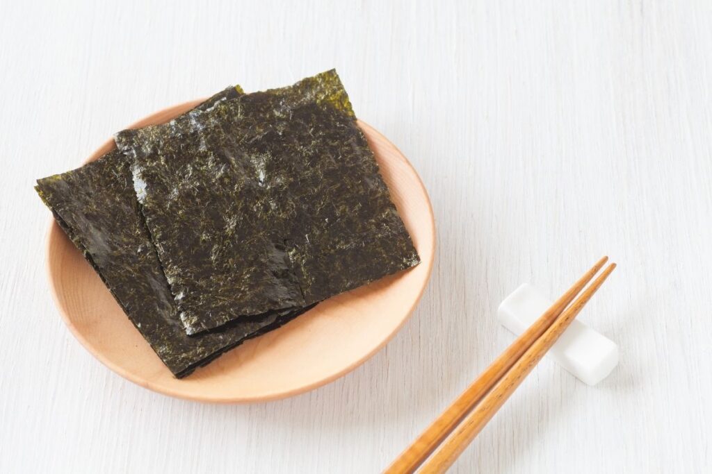 A plate of dark green nori sheets is arranged neatly, accompanied by a pair of wooden chopsticks. The simple and minimalist presentation emphasizes the texture of the nori, set against a light background.