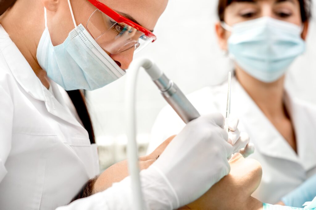 Un professionnel dentaire travaille soigneusement sur les dents d'un patient tout en portant un équipement de protection, y compris un masque et des lunettes. Un autre membre du personnel observe à proximité, également masqué. L'environnement suggère un cadre de cabinet dentaire propre et stérile.  