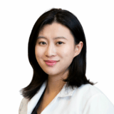 A smiling woman with shoulder-length black hair wears a white lab coat. She appears friendly and approachable, suggesting a professional setting, likely in healthcare or dentistry.