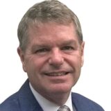 A smiling man with short gray hair and a business suit poses against a white background, conveying a professional and friendly demeanor.