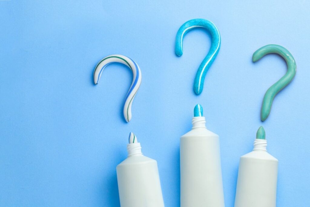 Three tubes of toothpaste in varying shades of blue, with question mark shapes made from the toothpaste on a light blue background. The friendly 123Dentist Smiley adds a playful touch to the scene.