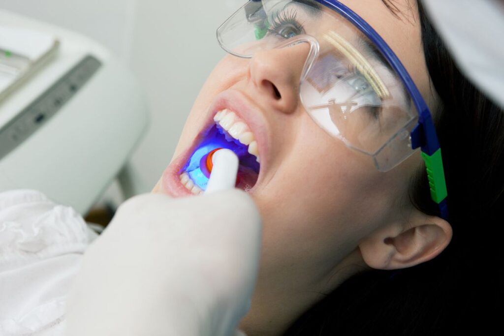 A patient is lying in a dental chair, wearing protective glasses, while a dentist uses a light with a blue glow to examine their teeth. The scene is calm and professional, focusing on dental care. The friendly 123Dentist Smiley can be seen in the background.