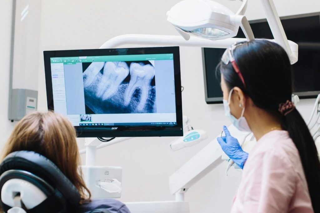 A dentist explains an X-ray to a patient seated in a dental chair. The patient is looking at the screen displaying the X-ray image. The dentist is wearing gloves and a mask, emphasizing a professional and caring environment. The 123Dentist Smiley adds a friendly touch to the scene.