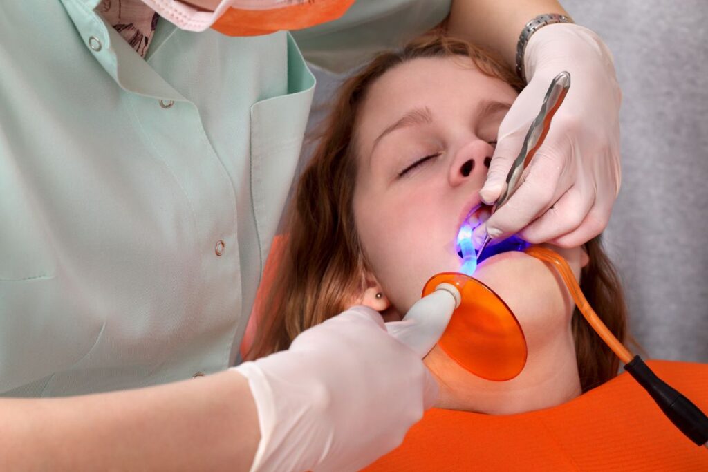 A dental professional treats a patient in a dental chair, using a tool with a blue light, while the patient relaxes with eyes closed. The scene captures a routine dental procedure, emphasizing care and comfort. The friendly 123Dentist Smiley is also present, adding a cheerful touch.
