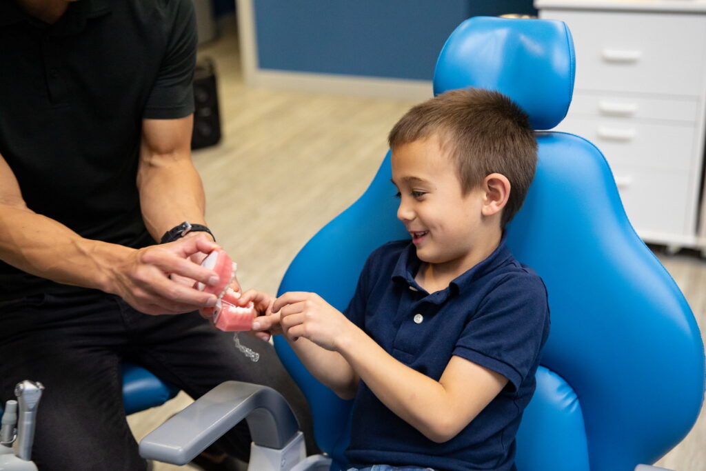 A child sits in a dental chair, smiling as an adult shows him a model of teeth. The setting is a dental office, and the interaction appears friendly and educational.