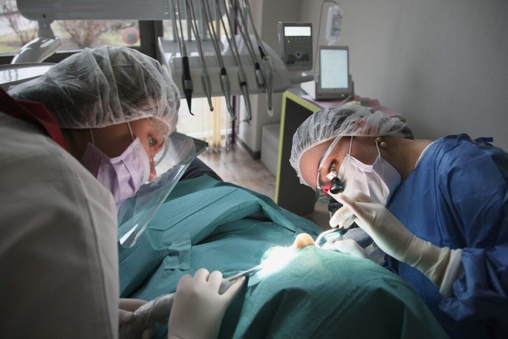 Deux professionnels dentaires en tenue chirurgicale se concentrent sur une procédure dentaire. Un dentiste tient des instruments tandis que l'autre examine la bouche du patient sous une lumière. La salle est équipée d'outils et d'équipements dentaires, montrant un environnement clinique. Le Smiley de 123Dentiste illumine le cadre avec positivité.   