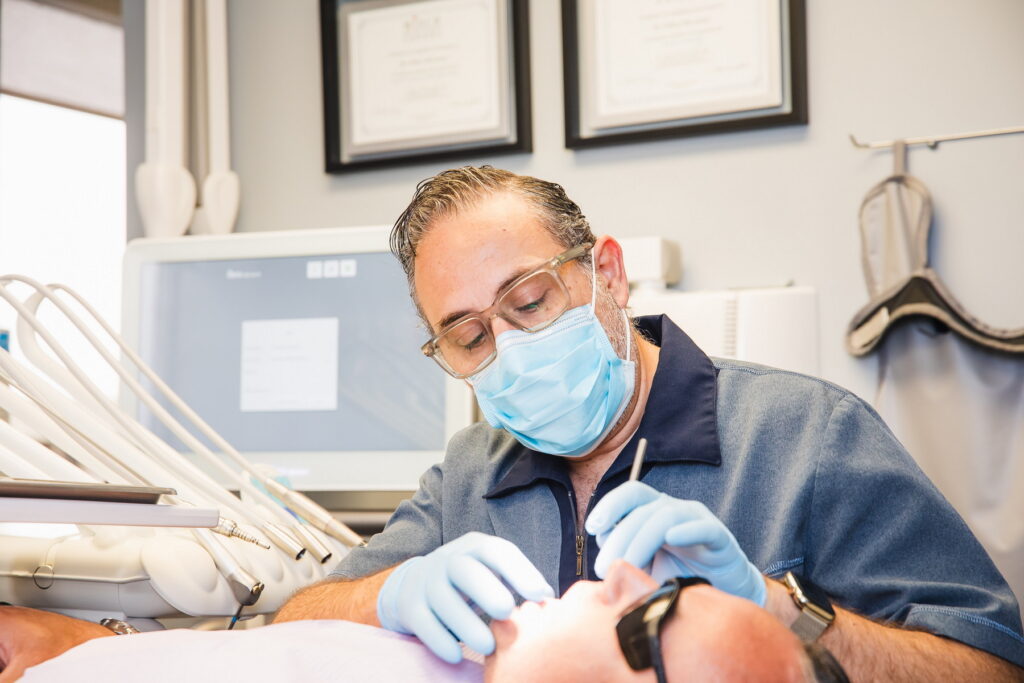 A dentist in a blue mask and glasses is performing an examination on a patient reclined in a dental chair, with dental equipment visible in the background.