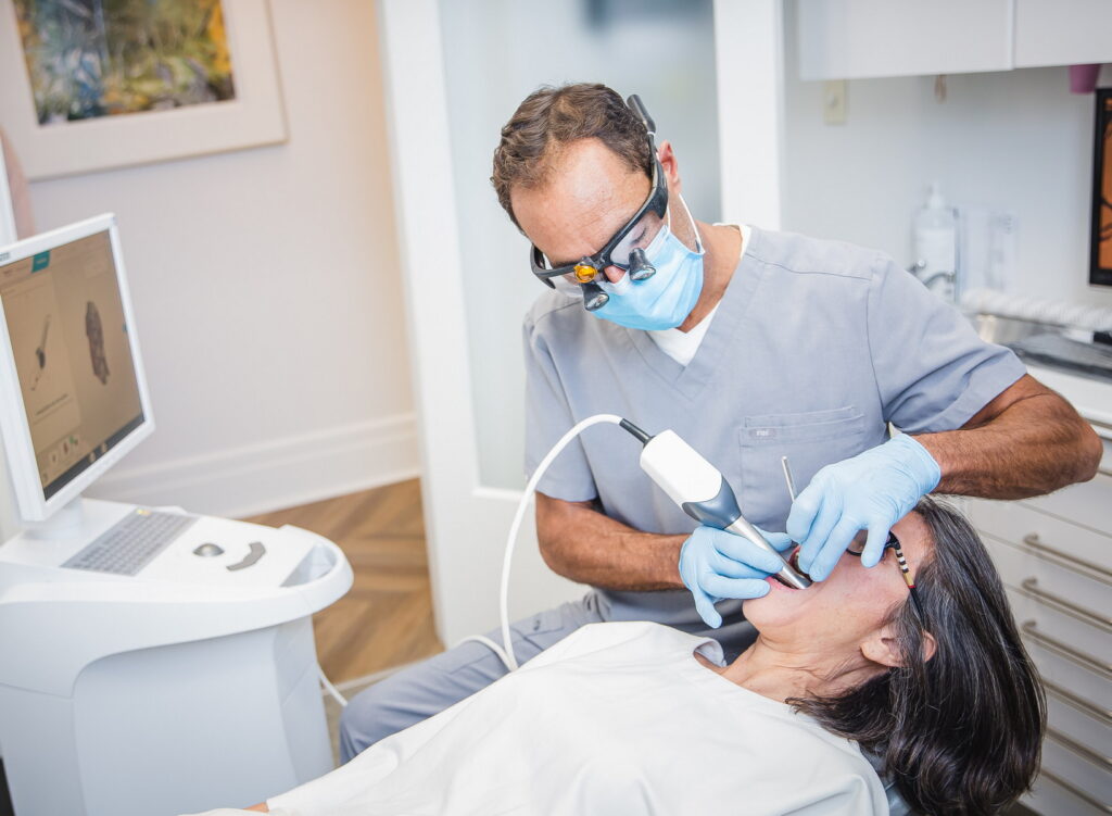 A dentist in scrubs is examining a patient's mouth using a dental device while the patient is seated in a dental chair. A computer screen is visible in the background, featuring the friendly 123Dentist Smiley.