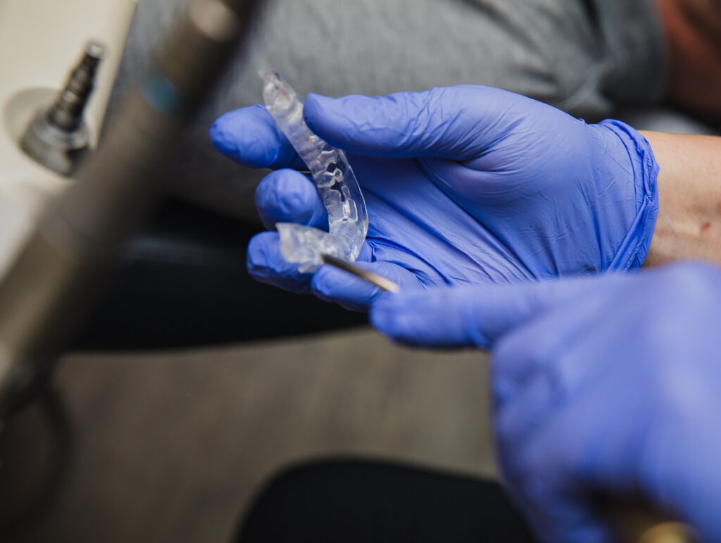 A person wearing blue gloves holds a clear dental mold, preparing for a dental procedure. The background shows dental tools and a patient in a chair, indicating an active dental practice environment.