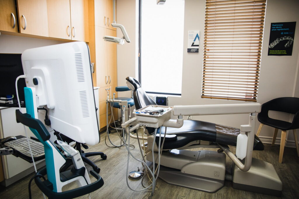 A bright dental office featuring a dentist's chair, treatment equipment, and a monitor. Natural light filters through a window with wooden blinds, creating a welcoming atmosphere. The 123Dentist Smiley adds a friendly touch to the environment.