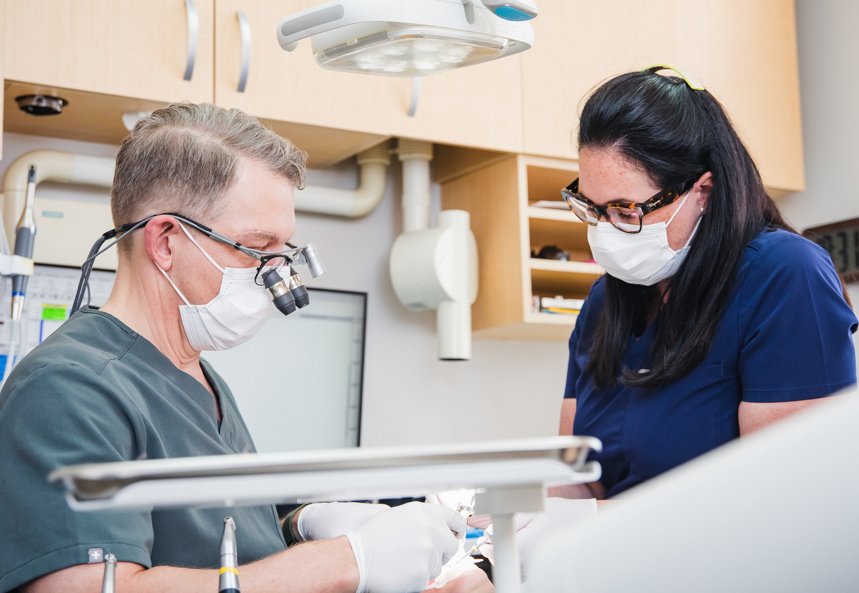 A dental professional wearing magnifying glasses collaborates with an assistant in a clinic. Both are wearing masks, focusing on a patient care procedure. The atmosphere reflects a clean, efficient dental environment.