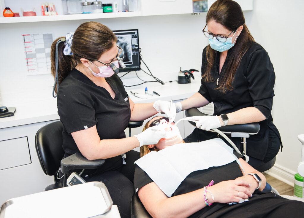 Two dental professionals in black scrubs, wearing masks and gloves, are working on a patient's teeth in a modern dental office. The patient is reclining in a dental chair with a protective bib. Tools and equipment are visible in the background.