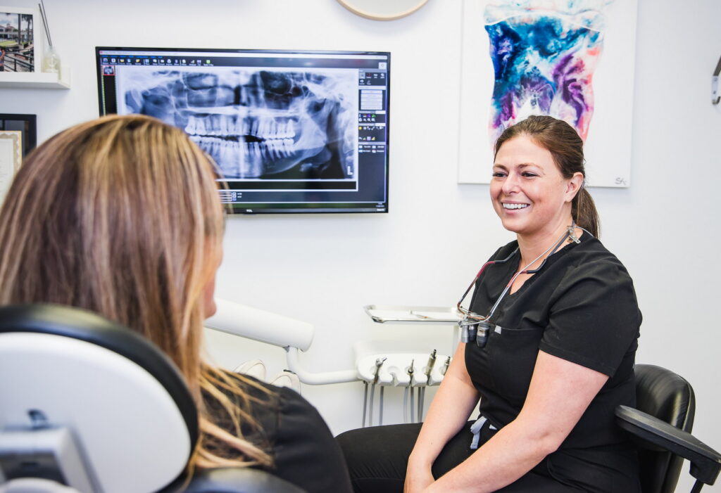 A smiling dental professional engages with a patient in a bright, modern clinic. An X-ray image is displayed on a screen behind them, reflecting a welcoming and friendly atmosphere during the dental visit. The 123Dentist Smiley adds a cheerful touch to the scene.