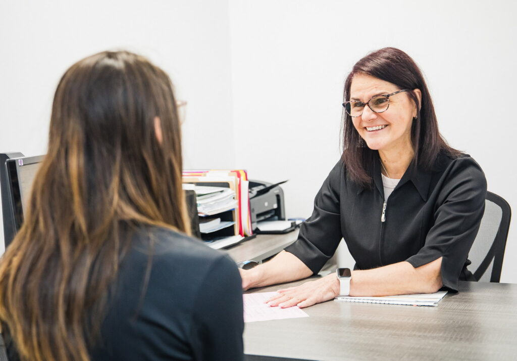 A smiling receptionist in a black outfit engages warmly with a client at a desk, surrounded by office materials. The interaction appears friendly and welcoming, suggesting a professional environment focused on client care.