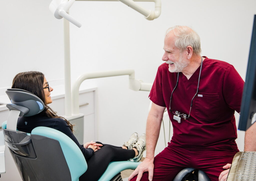 A dentist in a red scrub suit talks to a patient sitting in a dental chair. The patient, wearing glasses, looks relaxed and engaged in conversation. The clinical setting includes dental equipment and bright lighting.
