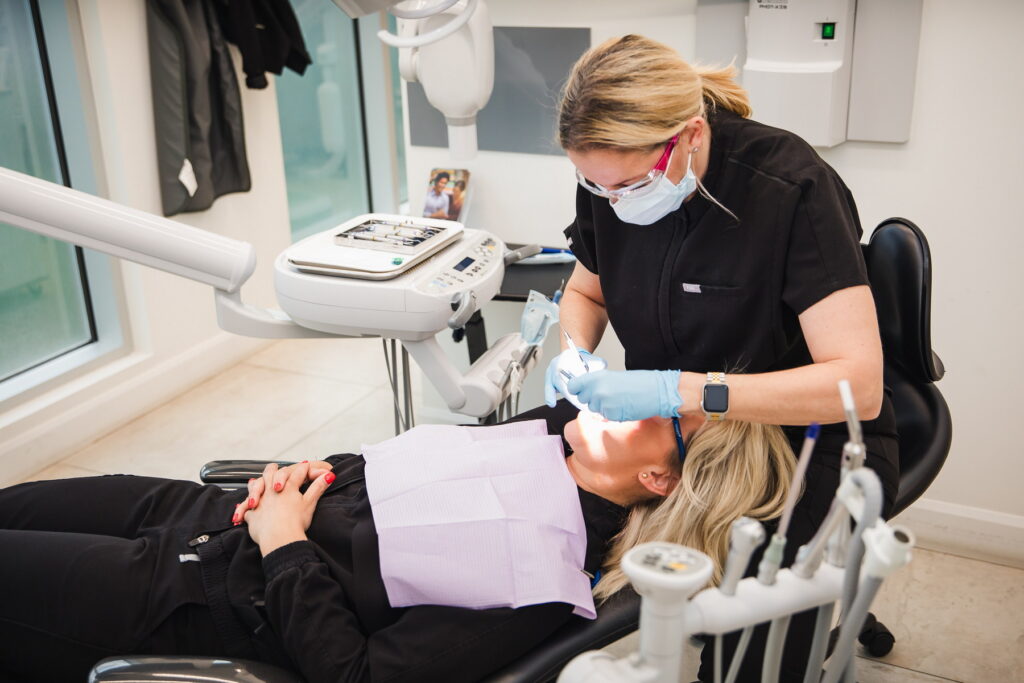 A dentist in protective gear is examining a patient reclining in a dental chair. The patient is relaxed with a dental bib on, while the dental office is bright and modern. The scene captures a typical dental check-up experience.