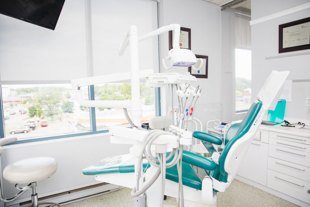 A modern dental office featuring a dental chair with teal accents, various dental instruments, and a bright window letting in natural light. The room is clean and organized, suitable for patient treatment. The friendly 123Dentist Smiley adds a cheerful touch to the environment.