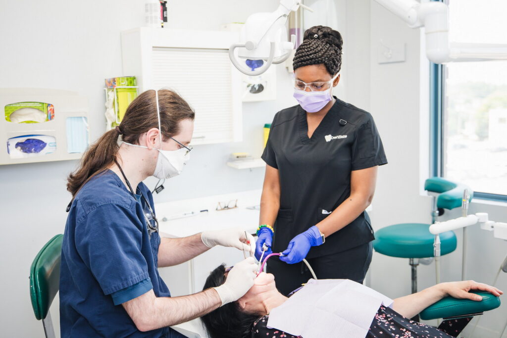 A dental professional and a dental hygienist are assisting a patient in a bright, modern dental clinic. The hygienist is wearing gloves and a mask while working on the patient's teeth, who is reclined in the dental chair. The atmosphere is clean and professional.