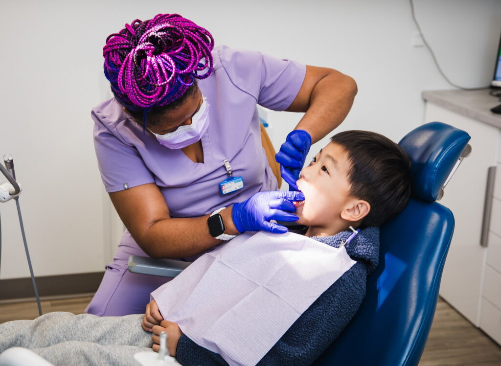 A dental professional in purple scrubs is examining a child's teeth in a dental office. The child is sitting in a dental chair, wearing a bib and looking up with his mouth open, while the professional uses dental tools to provide care.