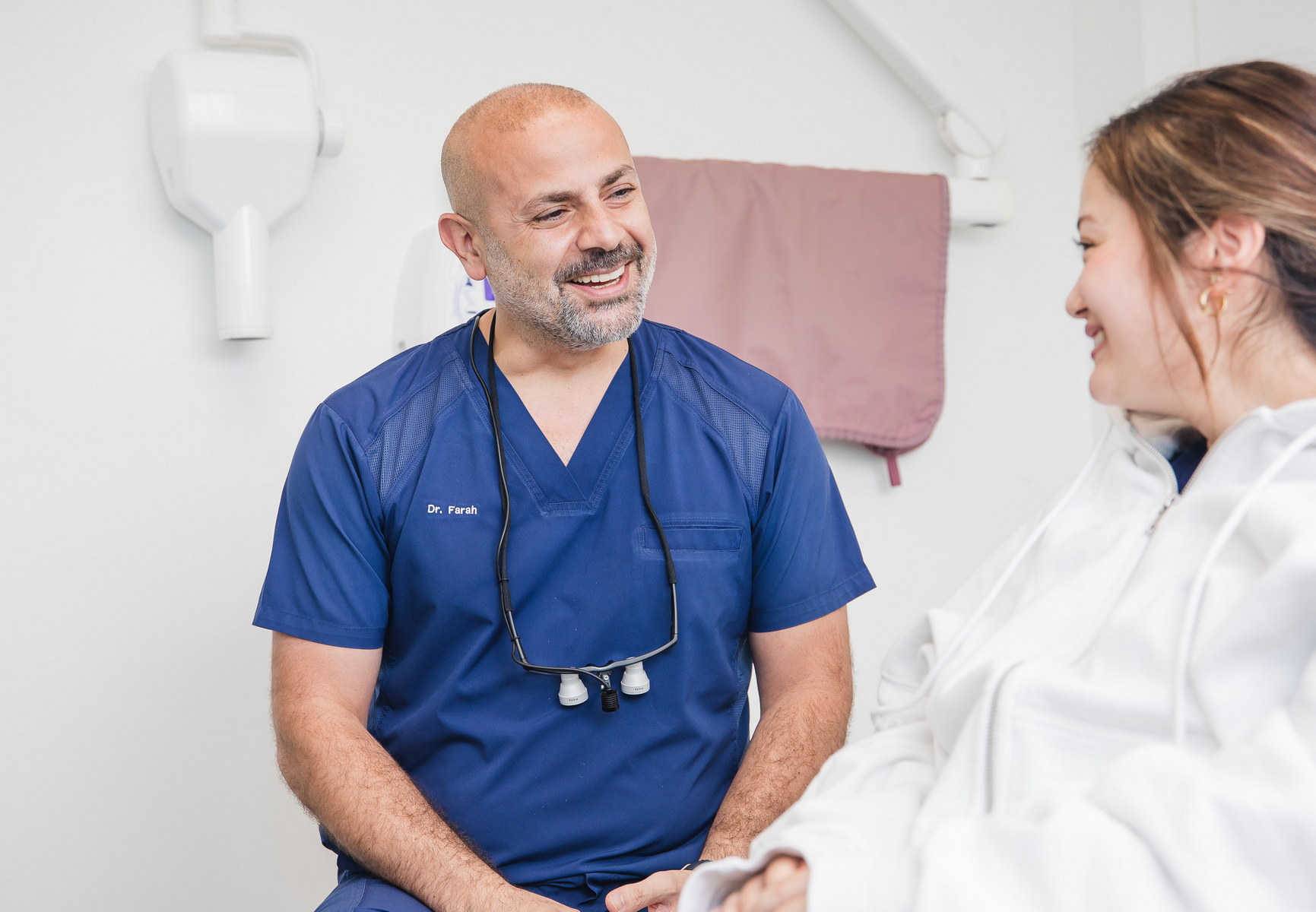 Un dentiste sympathique en tenue bleue sourit à un patient lors d'une consultation dans un cabinet dentaire lumineux. Le patient, vêtu d'un sweat à capuche blanc, semble détendu et engagé dans la conversation, créant une atmosphère accueillante pour les soins dentaires.
