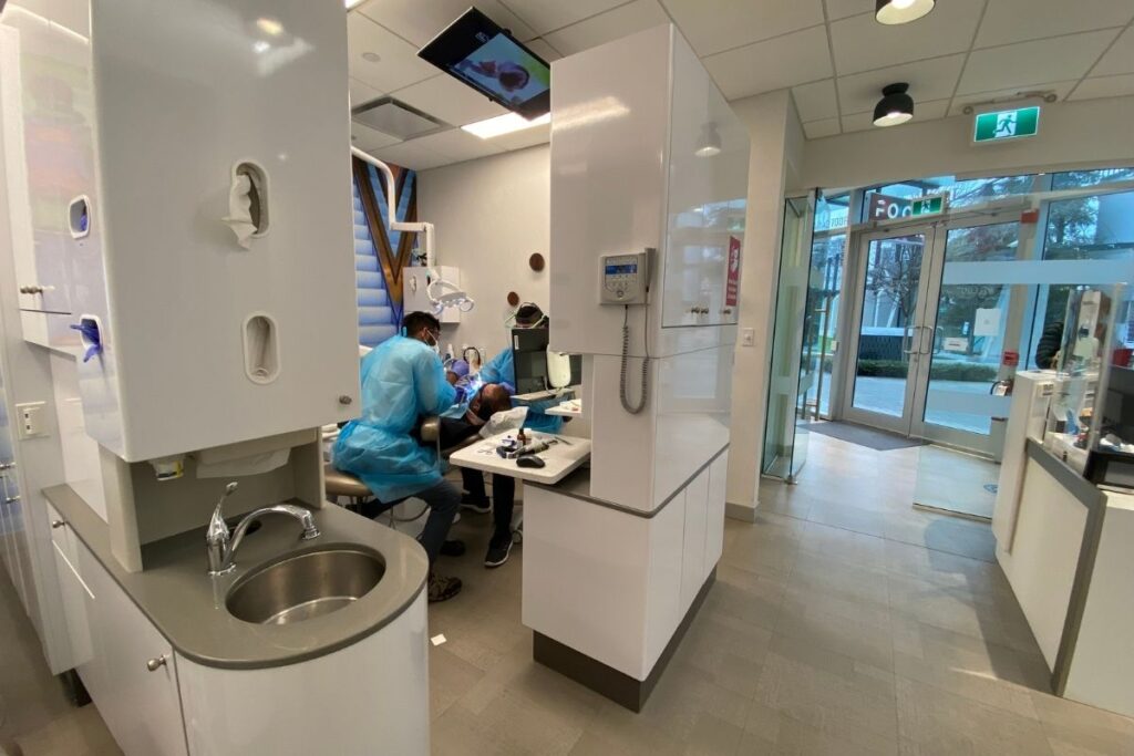 A bright and modern dental office is shown, featuring dental staff working with a patient. The space is clean and well-organized, with dental equipment visible. Natural light streams in through large windows, creating a welcoming environment. The 123Dentist Smiley adds a friendly touch to the atmosphere.