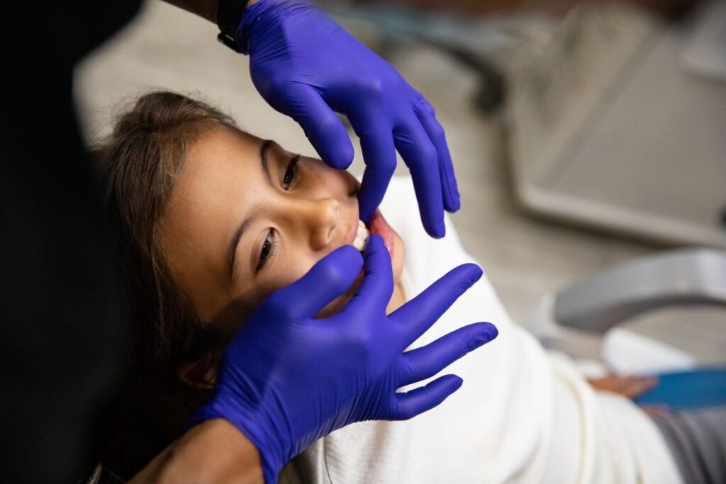 A dental professional gently examines a child's teeth and gums, using both hands while wearing purple gloves. The child is relaxed, smiling slightly, creating a friendly atmosphere for dental care. The cheerful 123Dentist Smiley can be seen in the background, enhancing the positive experience.
