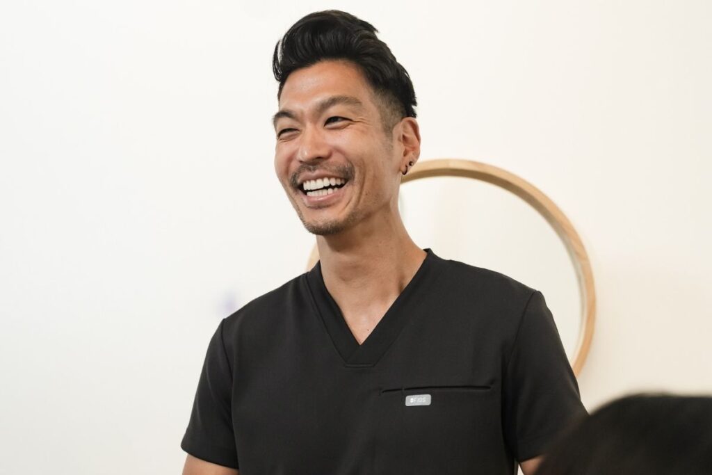 A smiling man in a black medical scrubs stands in a bright room, exuding warmth and approachability. He appears engaged in conversation, radiating positivity and friendliness.