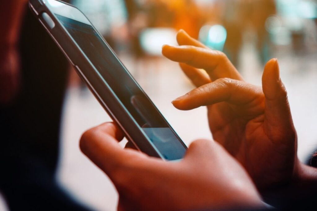 A close-up of a person's hands interacting with a smartphone, focusing on the screen while in a bustling environment. The image highlights technology usage in daily life.