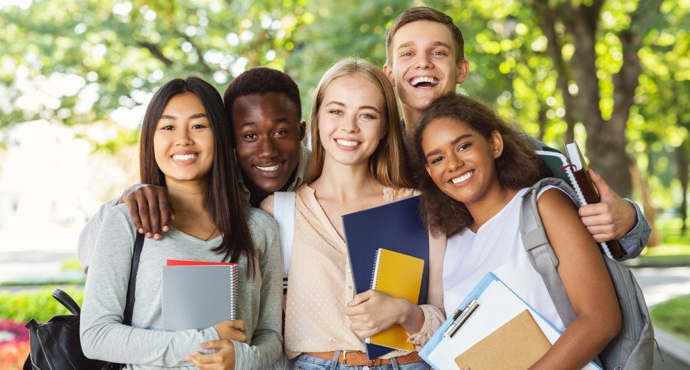 A diverse group of five smiling students stands together outdoors, holding books and notebooks. They appear joyful and engaged, showcasing a friendly and supportive environment, with trees in the background. The scene conveys camaraderie and positivity among friends.