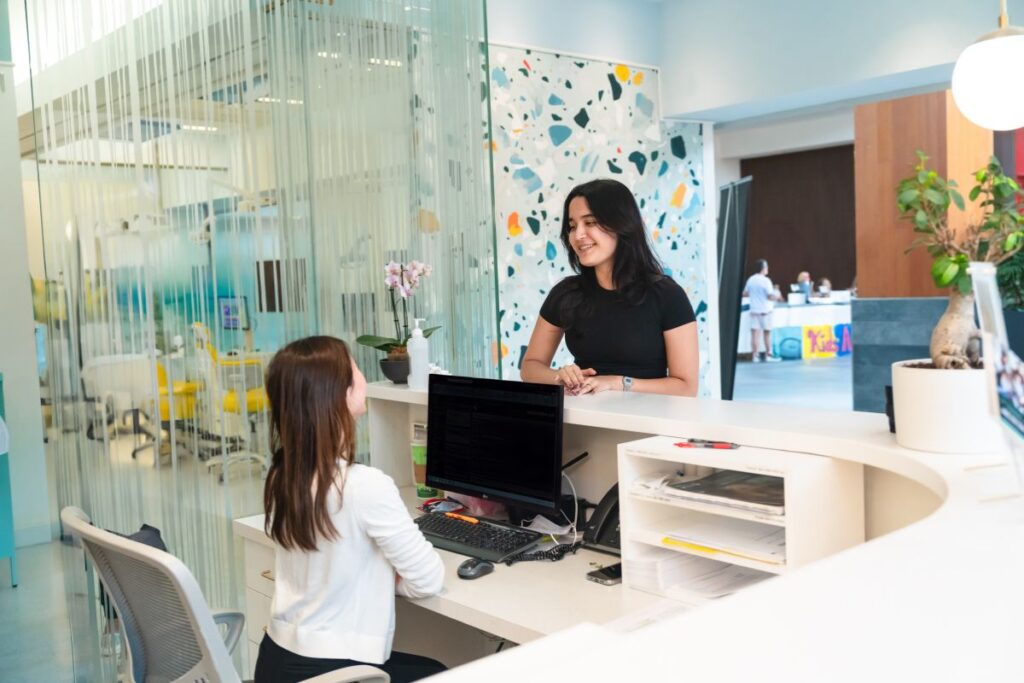 A reception area in a dental office features two smiling women, one seated at a desk with a computer and the other engaging in conversation from behind the counter. Colorful decor and plants add a welcoming atmosphere. The friendly 123Dentist Smiley can be seen nearby.