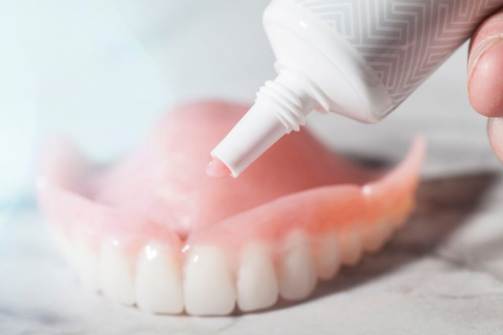 A hand is holding a tube of dental cream above a pair of dentures, highlighting dental care and hygiene.