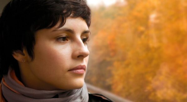A young woman gazes thoughtfully out of a window, with a backdrop of warm autumn trees. Her short dark hair frames her serene expression, while she wears a cozy scarf, embodying a moment of reflection and calm.
