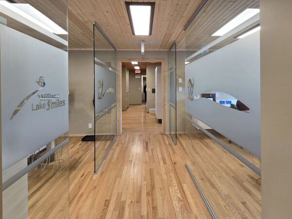 A modern dental office hallway featuring light-colored wooden floors, frosted glass doors with elegant signage, and soft lighting creating a welcoming atmosphere. The friendly 123Dentist Smiley adds a cheerful touch to the space, enhancing the inviting environment for patients.