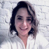 A woman with curly hair smiles warmly at the camera, wearing a white blouse. She stands in front of a textured white brick wall, exuding a welcoming and friendly vibe.